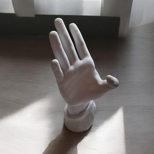 May include: White ceramic hand sculpture with an open palm, the fingers are extended and the thumb is slightly bent. The sculpture is on a light wood surface.