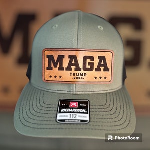 May include: A green and black trucker hat with a brown leather patch that says "MAGA TRUMP -2024-." The patch has four stars on each side. The hat has a black mesh back and a Richardson 112 adjustable strap.