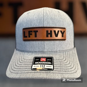 May include: A gray and black trucker hat with a brown leather patch that says "LFT HVY". The hat has a black bill and a black adjustable strap with a red "R" logo and the text "EST. 1970 RICHARDSON. ORIGINAL 112 TRUCKER ADJUSTABLE".