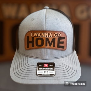 Peut inclure: Un chapeau de camionneur gris et noir avec un patch en cuir marron qui dit "I Wanna Go Home". Le chapeau a un dos en maille noire et une visière noire. Le chapeau a également une étiquette noire et rouge qui dit "Richardson" et "112".
