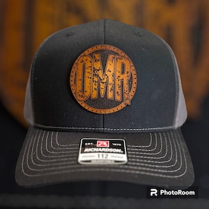 Może przedstawiać: Czarna i szara czapka trucker z brązową skórzaną naszywką z inicjałami "OMR" w stylu vintage.