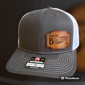 May include: A gray and white trucker hat with a brown leather patch that says "Sounds Like Bullshit To Me". The hat is adjustable and has a Richardson 112 label.