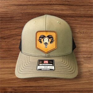 Peut inclure: Un chapeau de camionneur vert et noir avec un patch en cuir marron représentant un crâne de bélier blanc avec des cornes noires. Le patch a la forme d'un bouclier et un fond jaune. Le chapeau porte l'inscription "EST. 1970 RICHARDSON." sur le devant.
