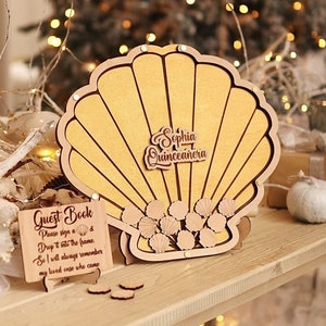 May include: A wooden seashell-shaped guest book frame with the text "Sophia Quinceañera." The frame has a light brown colour and includes small wooden seashell accents. A small sign reads "Guest Book" with instructions.