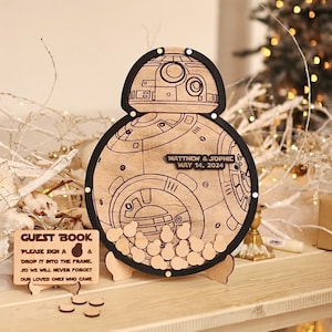May include: A wooden BB-8 guest book with a black frame and a sign that reads "GUEST BOOK PLEASE SIGN A & DROP IT INTO THE FRAME. SO WE WILL NEVER FORGET OUR LOVED ONES WHO CAME". The BB-8 is decorated with small wooden circles and has a sign that reads "MATTHEW & SOPHIE MAY 14, 2024".