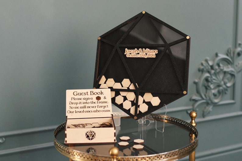 May include: A wooden guest book box with a hexagonal cutout and a sign that reads "Guest Book Please sign a & Drop it into the frame. So we will never forget Our loved ones who came". A black geometric frame with a clear window and the text "Joseph & Juliana October 12th 2024" is on a gold stand.