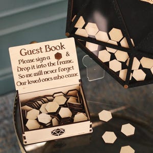 May include: A wooden guest book box with a sign that reads "Guest Book Please sign a & Drop it into the frame. So we will never forget Our loved ones who came". The box is open and filled with hexagonal wooden pieces. There are also some hexagonal pieces scattered on the table.