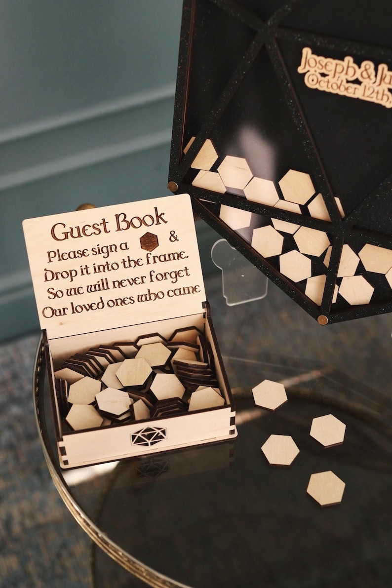 May include: A wooden guest book box with a sign that reads "Guest Book Please sign a & Drop it into the frame. So we will never forget Our loved ones who came". The box is open and filled with hexagonal wooden pieces. There are also some hexagonal pieces scattered on the table.