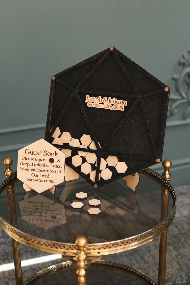 May include: A black geometric guest book frame with a wooden hexagon insert. The frame is on a gold stand and has the text "Joseph & Juliana October 12th, 2024" on the front. A separate wooden sign reads "Guest Book Please sign a & Drop it into the frame. So we will never forget Our loved ones who came"
