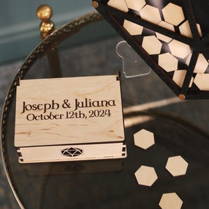 May include: A wooden box with a lid, engraved with the text "Joseph & Juliana October 12th, 2024". The box is sitting on a glass table with several wooden hexagonal pieces scattered around it.