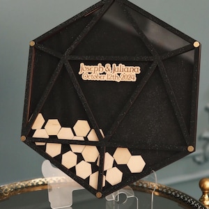 May include: Black geometric wedding guest book with a clear acrylic stand. The guest book is filled with wooden hexagonal pieces. The guest book has the text "Joseph & Juliana October 12th, 2024" on the front.
