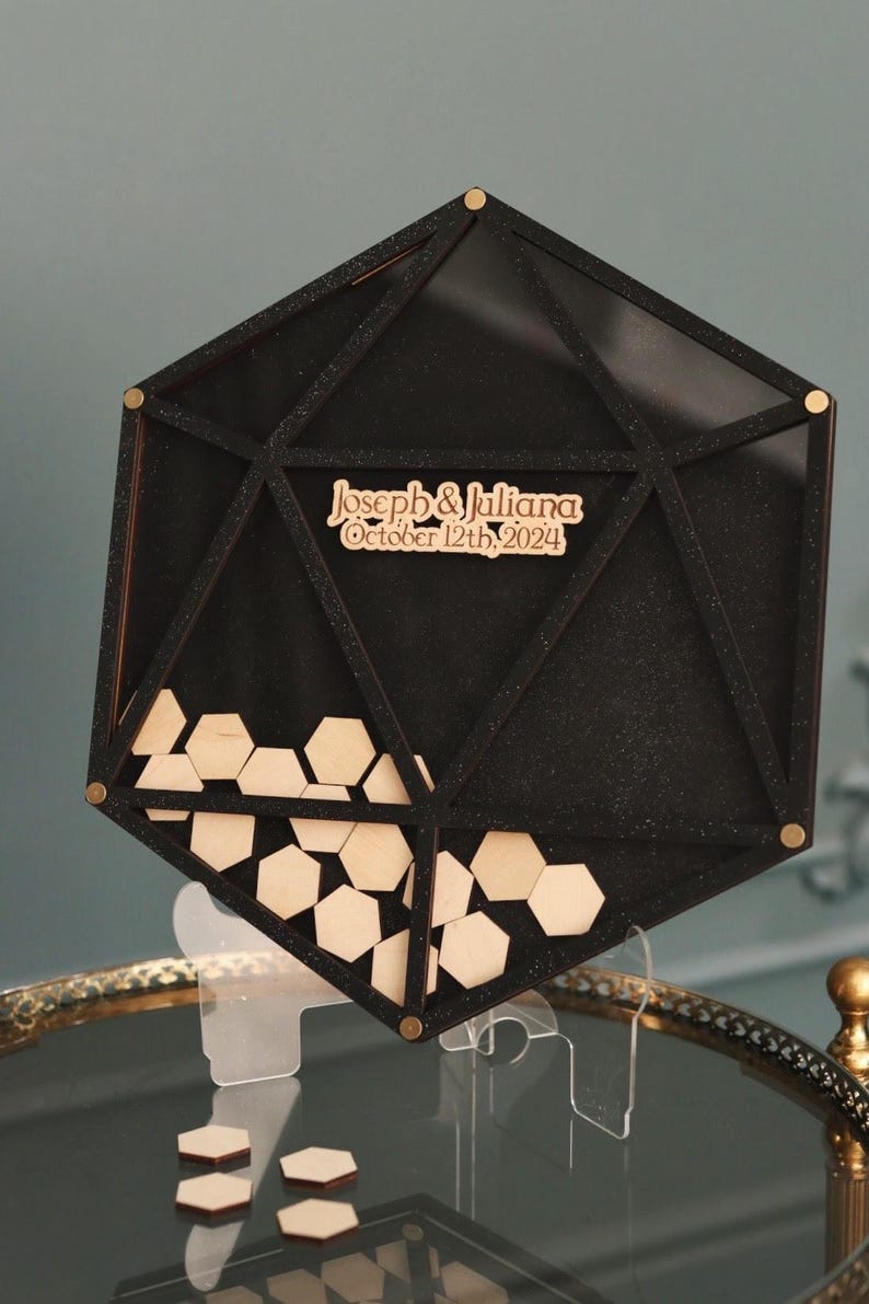 May include: Black geometric wedding guest book with a clear acrylic stand. The guest book is filled with wooden hexagonal pieces. The guest book has the text "Joseph & Juliana October 12th, 2024" on the front.