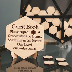 May include: A wooden guest book sign with the text "Guest Book" and instructions to sign a piece of wood and drop it into the frame. The sign is on a stand and there are several hexagonal pieces of wood on a table.