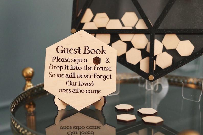 May include: A wooden guest book sign with the text "Guest Book" and instructions to sign a piece of wood and drop it into the frame. The sign is on a stand and there are several hexagonal pieces of wood on a table.