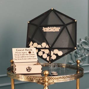 May include: A black wooden hexagonal guest book frame with a sign that reads "Please sign a & drop it into the frame. So one will never forget our loved ones who come."  The frame is on a gold metal stand with a round top and three legs.  The frame is filled with small wooden hexagonal pieces.