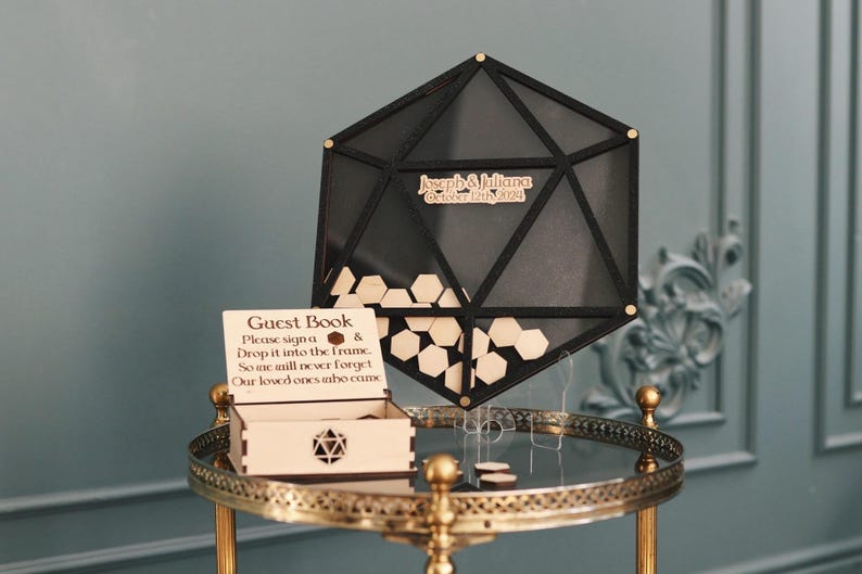 May include: A black wooden hexagonal guest book frame with a sign that reads "Please sign a & drop it into the frame. So one will never forget our loved ones who come."  The frame is on a gold metal stand with a round top and three legs.  The frame is filled with small wooden hexagonal pieces.