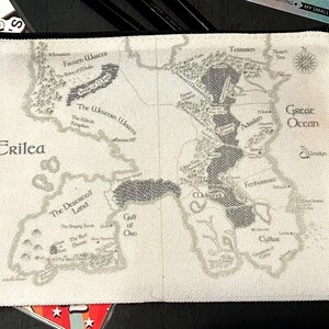 May include: A white canvas zipper pouch featuring a detailed map design in shades of gray. The map includes geographical features, place names, and the word "Crilea". The pouch has a black zipper and is likely used for storage.