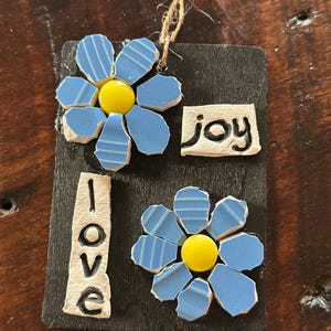 May include: A black wooden sign with two blue mosaic flowers and the words "love" and "joy" written in white.