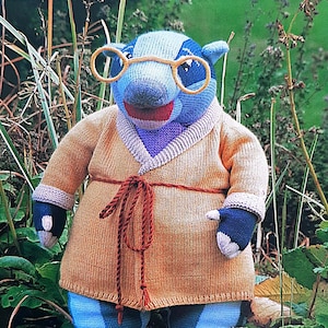 May include: A knitted plush toy animal wearing a brown robe with a yellow tie. The toy has a blue and purple body, a long snout, and round glasses.