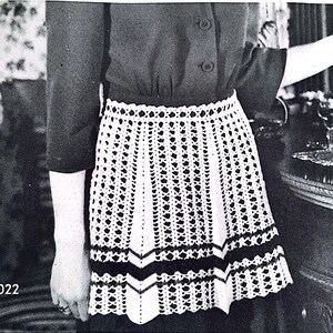 May include: A white crocheted apron with black trim. The apron is worn by a person and has a scalloped hem.