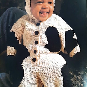 May include: A white knitted baby onesie with black cow spots and a hood. The onesie has a button fastening down the front. The baby is wearing the onesie and smiling.