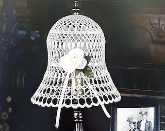 May include: A white crocheted lampshade with a white fabric flower on top. The lampshade is on a silver lamp base. The lamp is on a dark surface with a picture frame and a pocket watch.