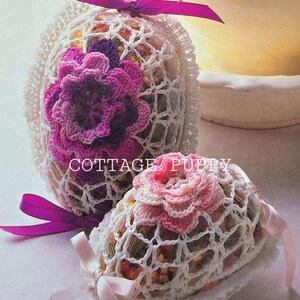May include: Two crocheted Easter eggs decorated with pink and purple flowers. The eggs are wrapped in white crocheted lace and tied with pink and purple ribbons. The text "COTTAGE PUPPY" is visible on the image.
