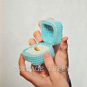 Puede incluir: Una caja de anillo de crochet azul claro con un anillo de oro en su interior. La caja está abierta y se sostiene en una mano. El texto "COTTAGE PUPPY" es visible debajo de la caja.
