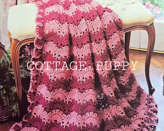 May include: A pink and burgundy crochet blanket with a scalloped edge. The blanket is draped over a white bench with a floral pattern on the wall behind it. The text "COTTAGE PUPPY" is visible on the blanket.