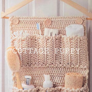 May include: A beige crocheted bathroom organiser with multiple pockets hangs on a wooden hanger. The organiser has buttons and is filled with toiletries, including a toothbrush, soap, and a washcloth. The text "COTTAGE PUPPY" is visible in the centre of the organiser.