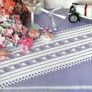 May include: White crocheted table runner with a delicate pattern of diamonds and stars. The runner is laid on a purple surface with a bouquet of pink and blue flowers, two martini glasses, and a toy car.