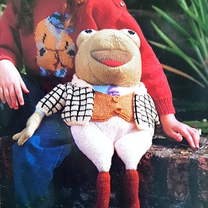May include: A knitted frog doll wearing a checkered vest and white pants. The frog has a brown and orange vest with a button and a purple bow tie. The frog is sitting on a brick wall with a child wearing a red sweater in the background.