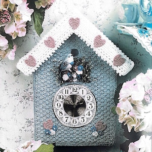 Puede incluir: Un reloj de ganchillo azul hecho a mano con forma de casa para pájaros, adornado con un tejado blanco y detalles de corazones rosas. Una pequeña figura de pájaro se encuentra encima de la esfera del reloj. El reloj está rodeado de flores rosas y blancas.