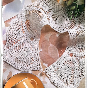 Puede incluir: Un collar de crochet blanco con un borde festoneado y una perla como adorno en el centro. El collar está colocado sobre una tela con estampado floral.