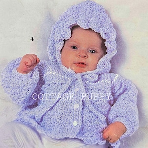 Puede incluir: Un bebé lleva una chaqueta de punto de ganchillo de color lavanda con ribetes blancos. La chaqueta tiene cierre de botón y un borde festoneado.