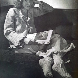 May include: A black and white photo of a woman wearing a crocheted cardigan and socks, sitting on a couch and reading a book.