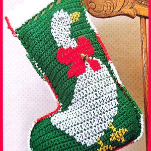 May include: A green and white crocheted Christmas stocking with a white goose design and a red bow with a bell. The stocking is hanging on a wooden chair.