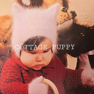 May include: A baby wearing a pink knitted cat ear hat and a red knitted sweater. The baby is holding a wooden toy. The image has the text "COTTAGE PUPPY" on it.