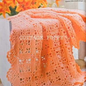 May include: A peach-coloured crocheted blanket with a scalloped edge. The blanket has a repeating square pattern and the text "COTTAGE PUPPY". The blanket is draped over a surface, with orange flowers in the background.