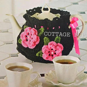 May include: A black crocheted teapot cosy with pink crocheted flowers and green leaves. The cosy is on a white teapot with a pink ribbon.