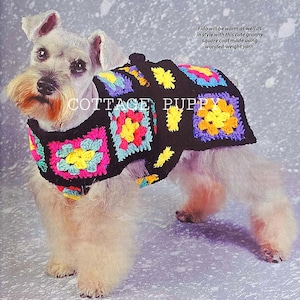 May include: A small, white dog wearing a black crocheted sweater with colorful granny squares. The sweater has a pom-pom on the collar. The text "COTTAGE PUPPY" is visible on the sweater. Fido will be warm as well as in style with this cute granny square coat made using worsted-weight yarn.