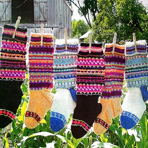 May include: Six colorful knitted socks hanging on a clothesline outdoors. The socks are striped with various colors, including pink, blue, green, yellow, and black. The socks are hanging against a backdrop of green foliage and a wooden barn.