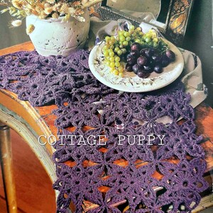 May include: A close-up of a purple crochet table runner with a floral design. The text "COTTAGE PUPPY" is visible. A plate of grapes and a vase of dried flowers are also present.
