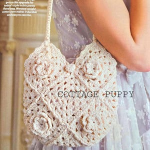 Puede incluir: Un bolso de mano blanco de ganchillo de forma granny square con un diseño floral. El bolso tiene una correa larga y está hecho de hilo de algodón de peso medio. El texto "COTTAGE PUPPY" es visible en el bolso.