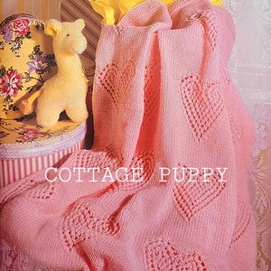 May include: A pink knitted baby blanket with a heart pattern. The blanket is folded and draped over a box with a stuffed giraffe toy on top. The text "COTTAGE PUPPY" is visible on the blanket.