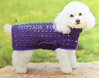 May include: A white dog wearing a purple crocheted sweater. The sweater has a cable knit pattern and is decorated with a small, white bow.