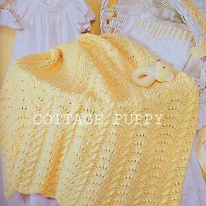 May include: A yellow knitted baby blanket with a delicate lace pattern. The blanket is draped over a white wicker basket with a white and yellow striped pillow inside. The text "COTTAG PUPPY" is visible on the blanket.