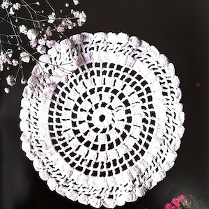 Könnte beinhalten: Weiße gehäkelte Deckchen mit einem runden Design und einem gewellten Rand. Das Deckchen liegt auf einem schwarzen Hintergrund mit weißen Blumen.