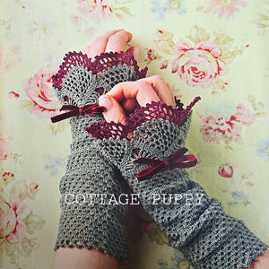 Op de afbeelding: Een paar grijze, vingerloze handschoenen van gehaakt garen met bordeauxrode kant en bordeauxrode satijnen strikken. De handschoenen liggen op een bloemige achtergrond.