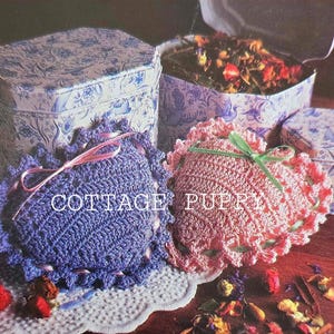 May include: Two crocheted heart-shaped pillows, one purple and one pink, with decorative ribbon accents. The pillows are sitting on a white doily with dried flower petals scattered around them.